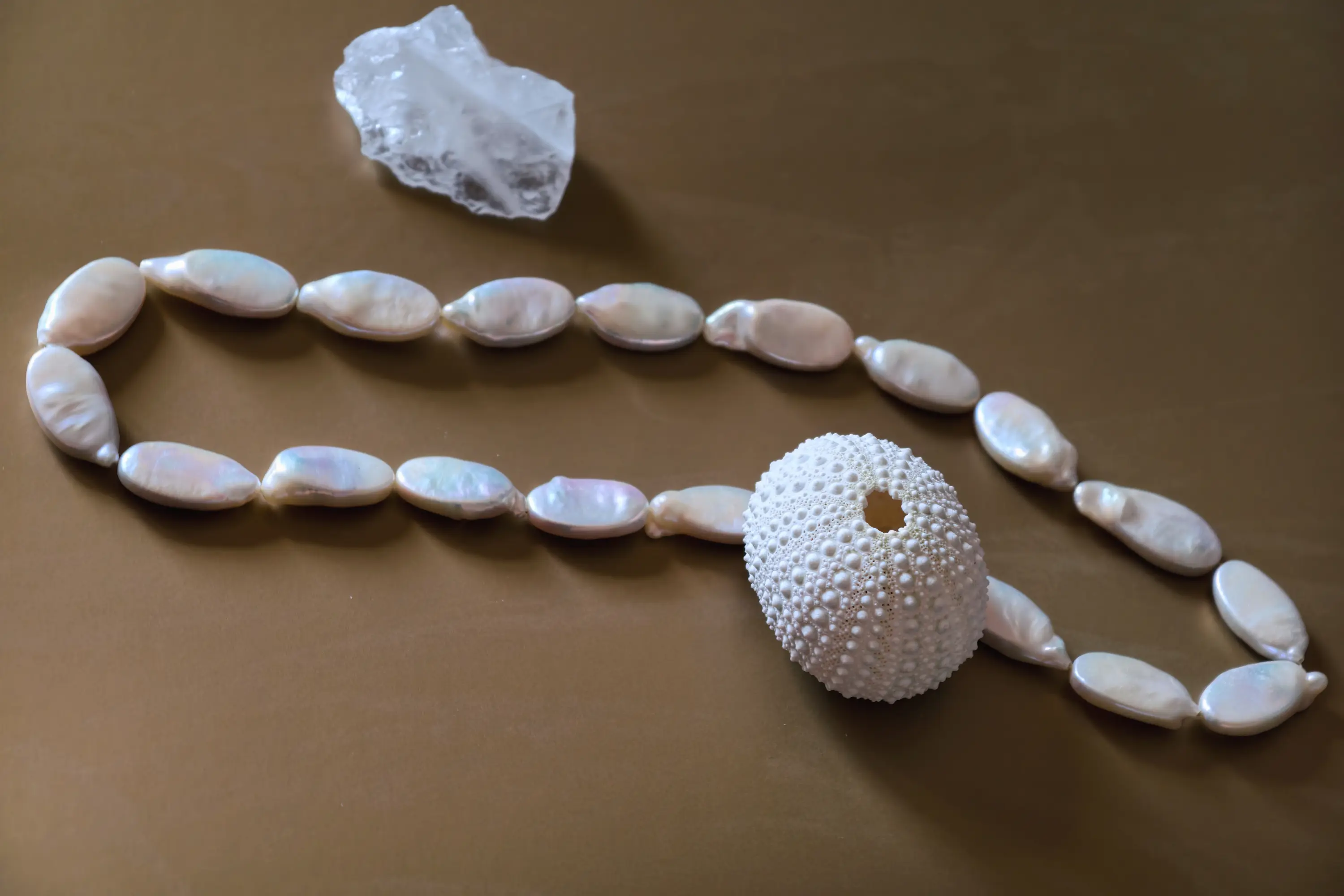 Elongated baroque pearl necklace arranged with a white sea urchin shell and a rough clear crystal on a warm brown surface.